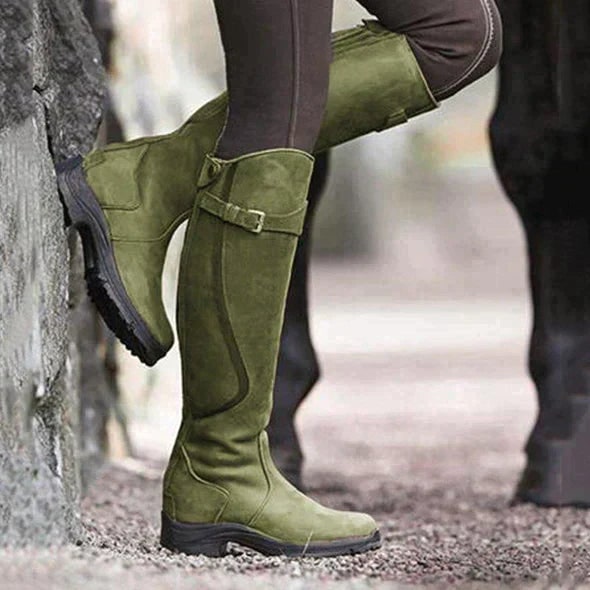 A person wearing green tall riding boots, leaning against a stone wall outdoors.