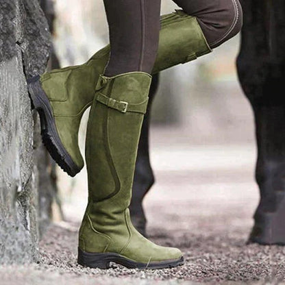 A person wearing green tall riding boots, leaning against a stone wall outdoors.