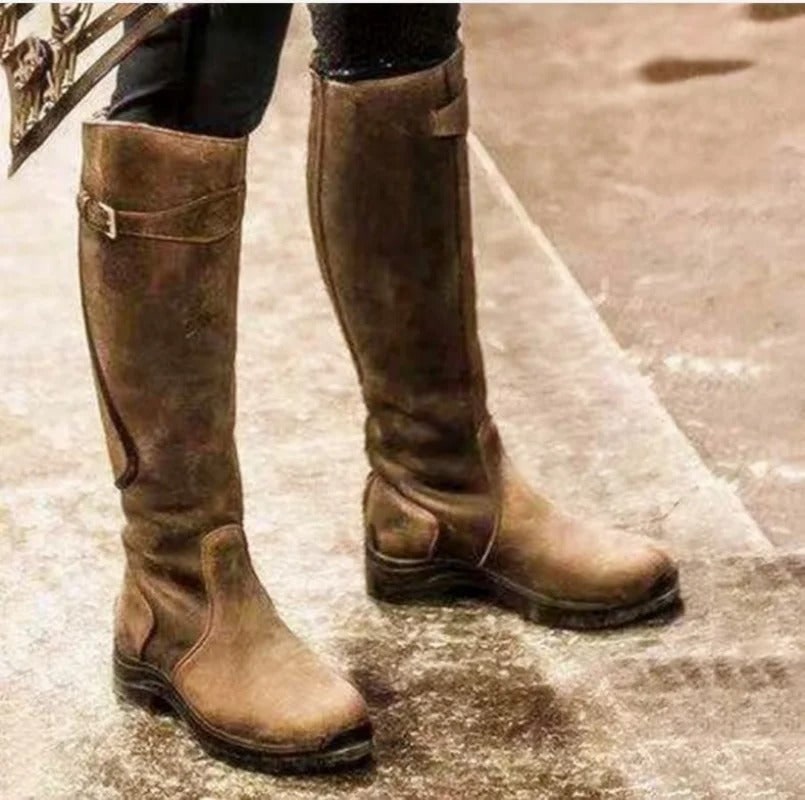 A person wearing brown tall riding boots, standing on a textured outdoor surface.