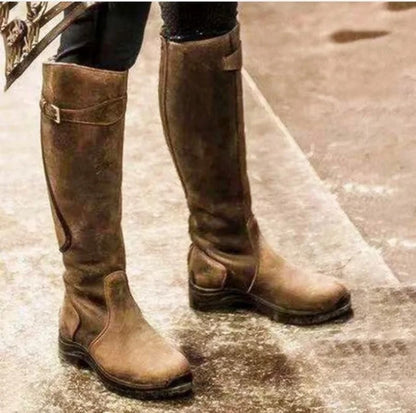 A person wearing brown tall riding boots, standing on a textured outdoor surface.