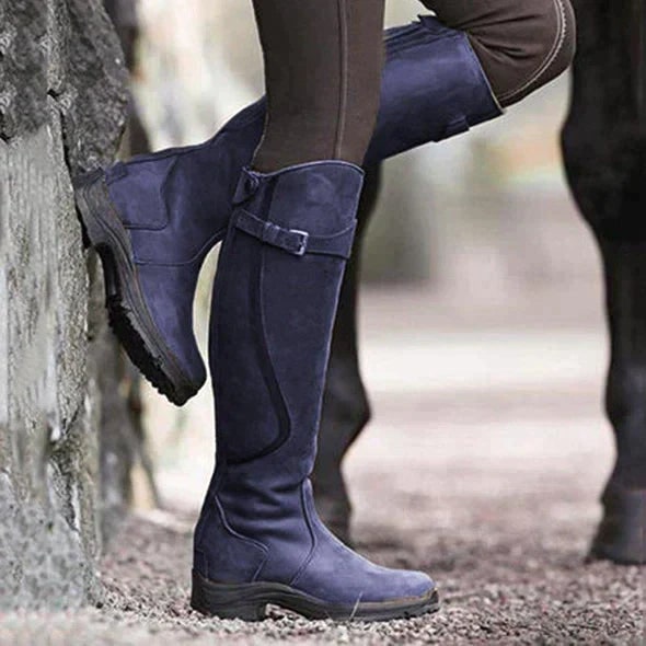 A person wearing blue tall riding boots, leaning against a stone wall outdoors.