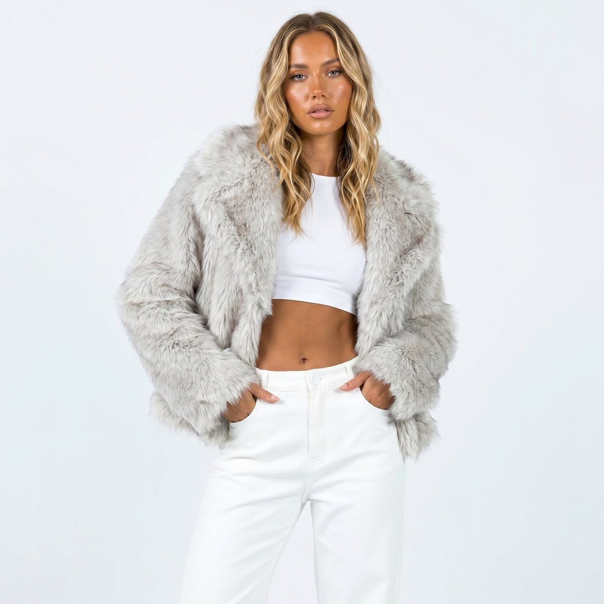Front view of a woman wearing a light grey cropped faux fur jacket over a white crop top and white pants.