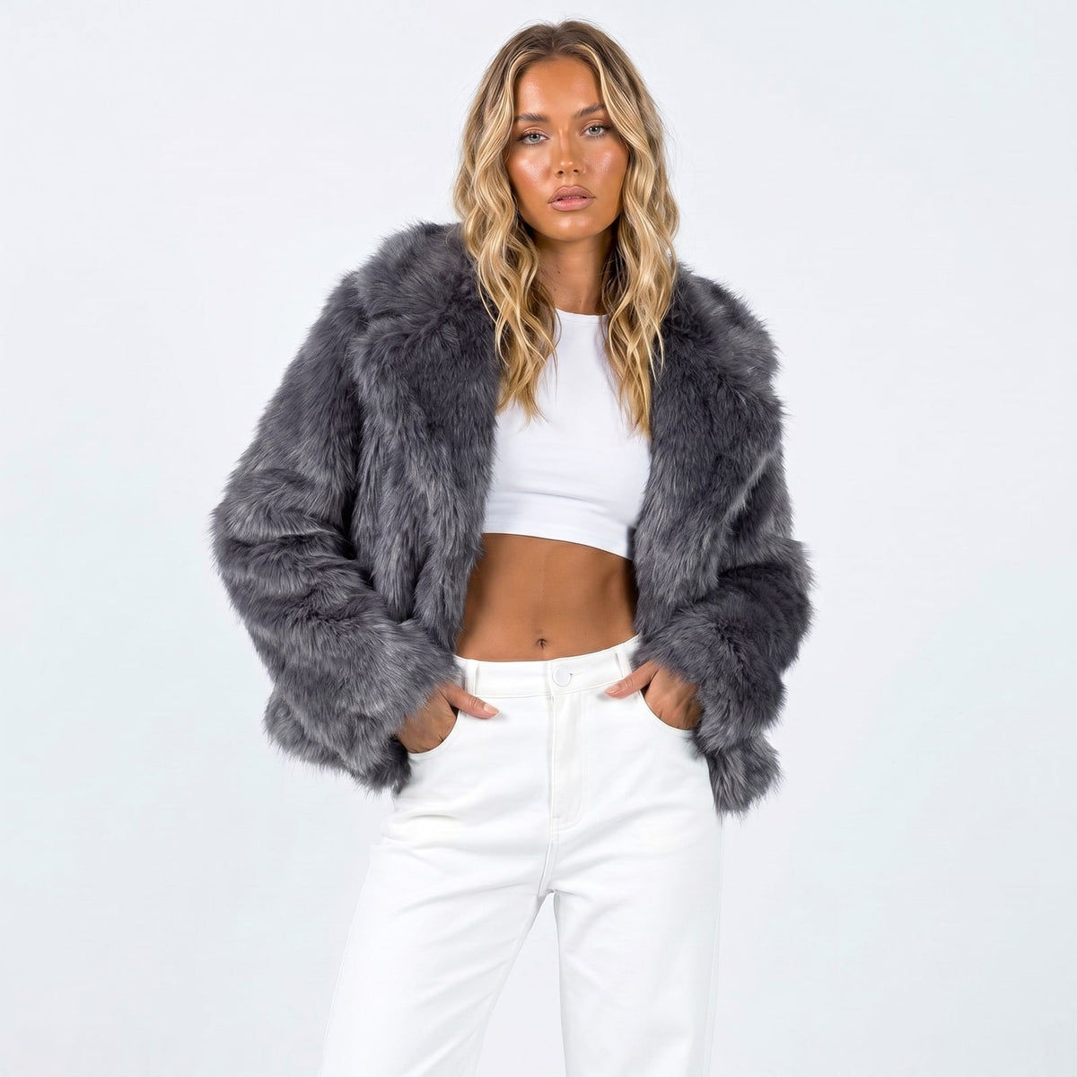 Front view of a woman wearing a dark grey cropped faux fur jacket over a white crop top and white pants.