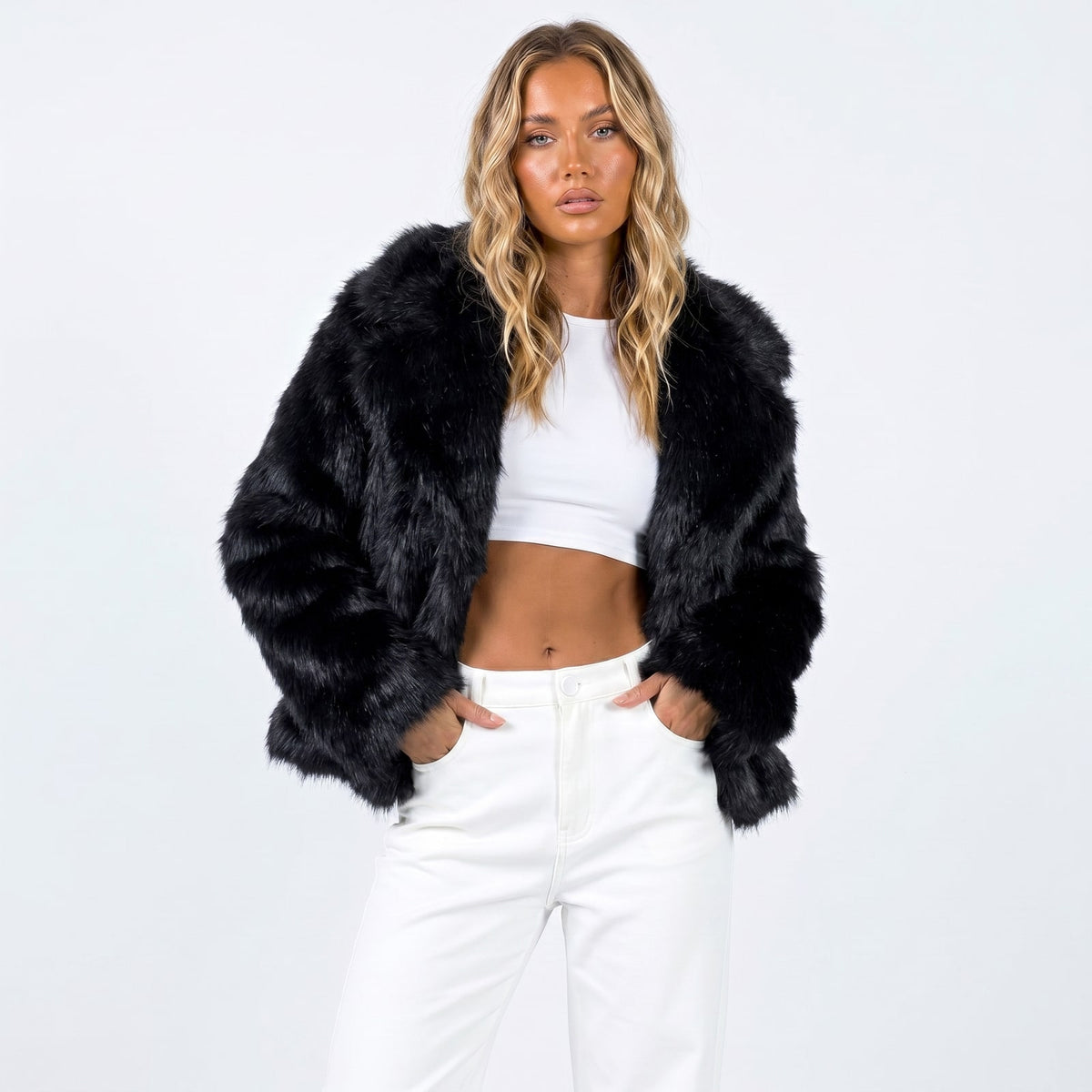 Front view of a woman wearing a black cropped faux fur jacket over a white crop top and white pants.