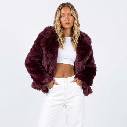A blonde woman models a burgundy faux fur jacket, white crop top, and white pants, posing with hands in pockets.