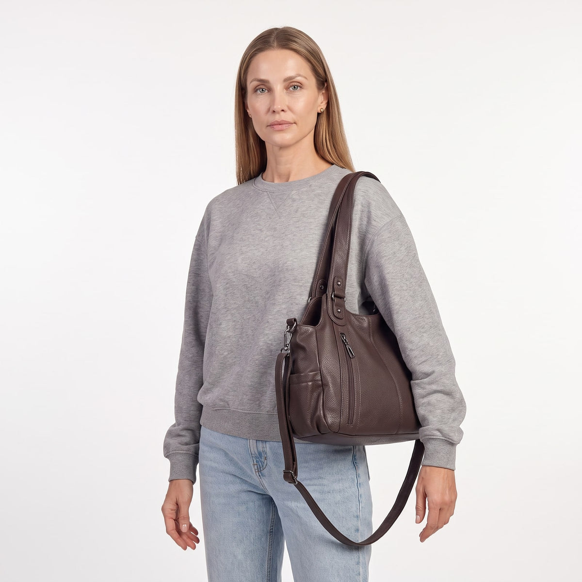 A woman wearing a grey sweatshirt and jeans carries the dark brown faux leather hobo bag over her shoulder, looking directly at the camera.