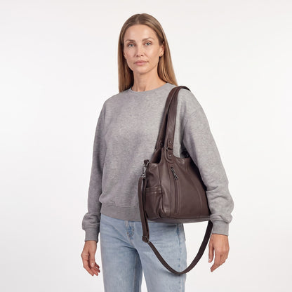A woman wearing a grey sweatshirt and jeans carries the dark brown faux leather hobo bag over her shoulder, looking directly at the camera.