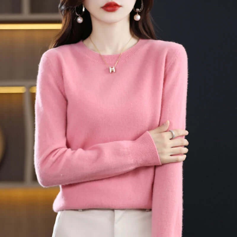 A model wears a pink long-sleeved crew neck sweater, accessorized with a gold 'H' pendant necklace.