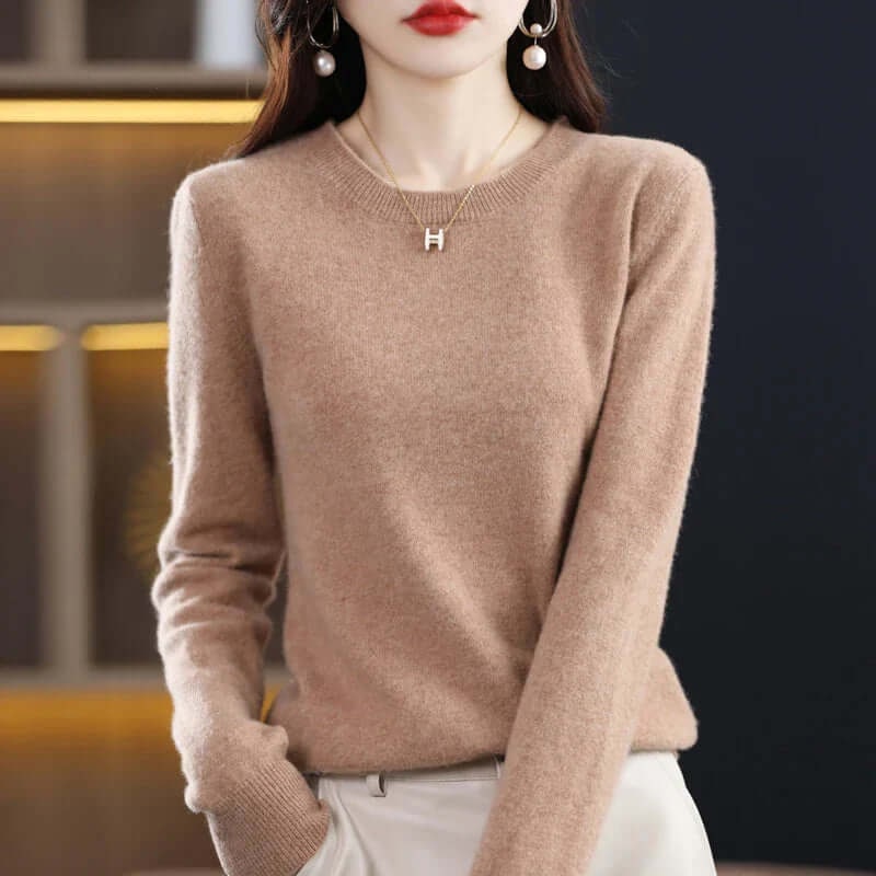 A model wears a light brown long-sleeved crew neck sweater, accessorized with a gold 'H' pendant necklace.
