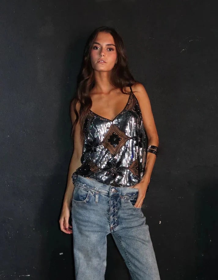 Full-body front view of a woman wearing a silver and black embellished sheer camisole top with light wash jeans, posing against a dark wall.