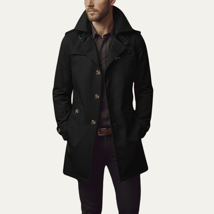 Front view of a man wearing a black single-breasted trench coat with a patterned shirt and dark trousers.