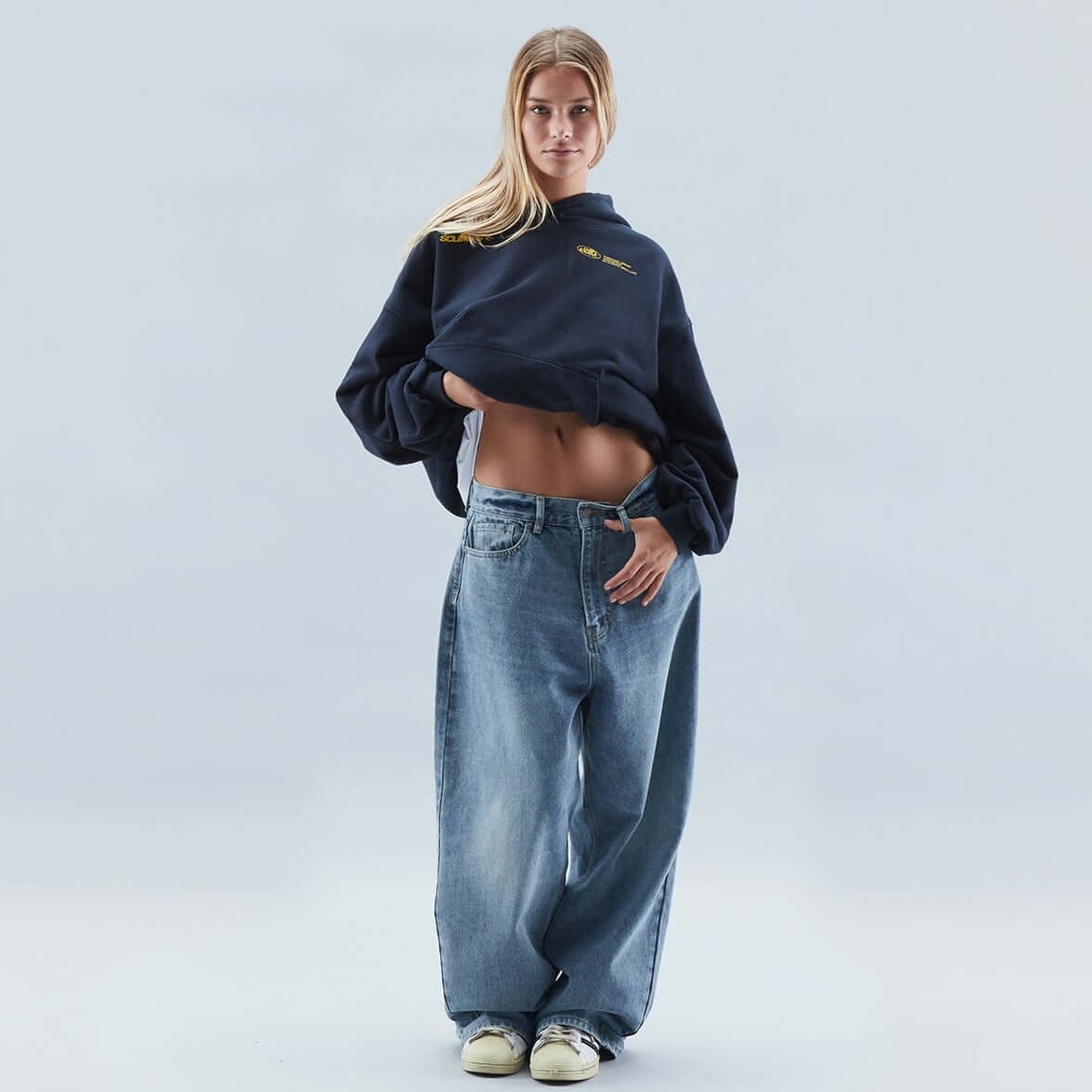 On-model front view of a dark blue oversized hoodie, with the model lifting the hem to reveal her midriff.