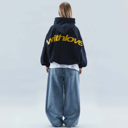 On-model back view of a dark blue oversized hoodie with a large yellow 'withlove' graphic, worn with light wash baggy jeans.